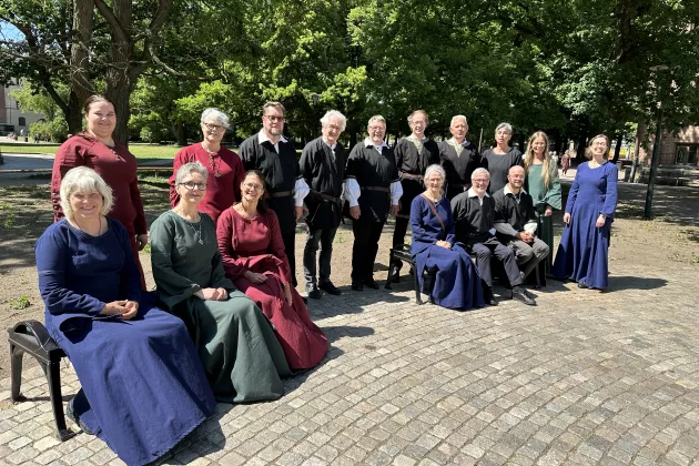 Kvinnor och män som står och sitter på bänkar, klädda i enkla medeltidsinspirerade kläder i färgerna blått, svart, mörkrött och grönt.