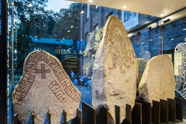 Tre runstenar står på rad mot en bakgrund av en glasruta. Den vänstra har en runrad och ett kors, den i mitten en stiliserad beriden figur, och den högra är inte ifylld med färg. Genom glaset syns lövverk i kvällningen.