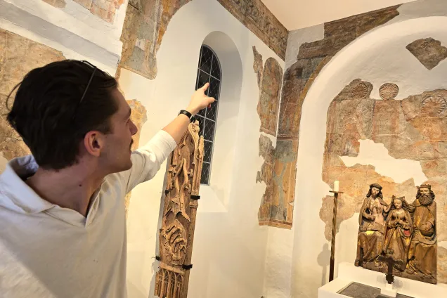 En manlig guide, snett vänd från kameran, i vit tröja stående till vänster i bild pekar uppåt i ett rum täckt av fragmenterade bruna medeltida kalkmålningar. Till vänster står medeltida träskulpturer. Till höger syns i rummet ett fönster och en dekorerad korstolsgavel i trä. 