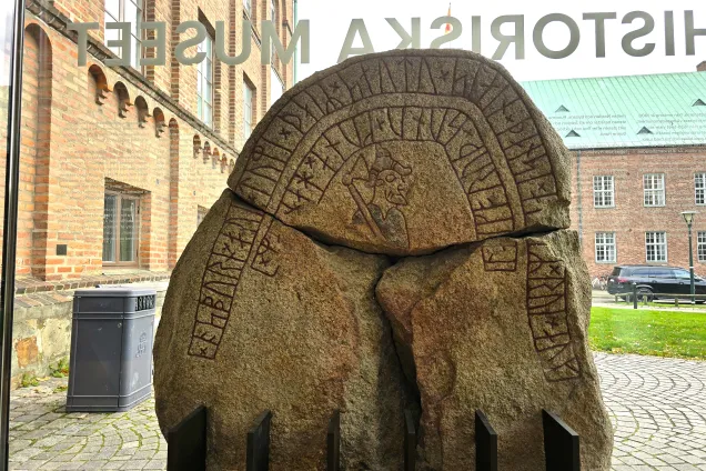 En stor runsten som är sprucken i 3 delar. En runrad går längs stenen runt en bild på en man med yxa.