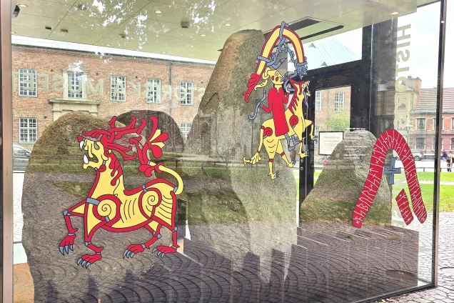 tre runstenar står bakom glas. På glaset finns bilder i framförallt rött och guld som visar hur stenarna har varit målade.