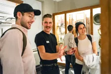 En guide i mörka kläder, vänt snett mot kameran, håller visning av en museiutställning. Till höger och vänster om honom står två leende åhörare och lyssnar, båda i ljusa kläder och ryggsäckar. I bakgrunden syns en annan besökare med ryggen vänd mot höga glaserade montrar som står mot väggarna fyllda med museiföremål. 