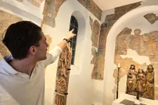 En manlig guide, snett vänd från kameran, i vit tröja stående till vänster i bild pekar uppåt i ett rum täckt av fragmenterade bruna medeltida kalkmålningar. Till vänster står medeltida träskulpturer. Till höger syns i rummet ett fönster och en dekorerad korstolsgavel i trä. 
