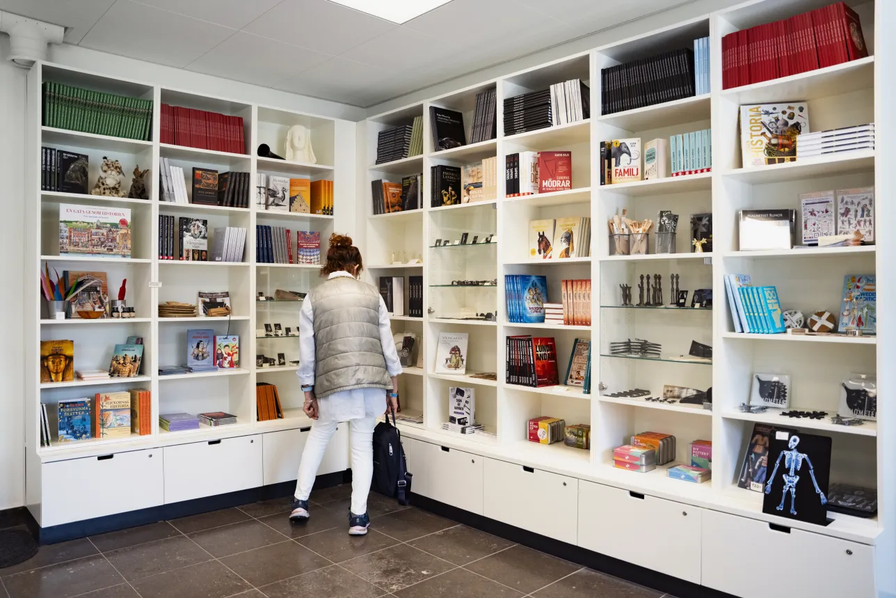 En kvinna i ljusa kläder står i en butik med vita hyllor. I hyllorna finns böcker, fjäderpennor, smycken och små bronsstatyetter.