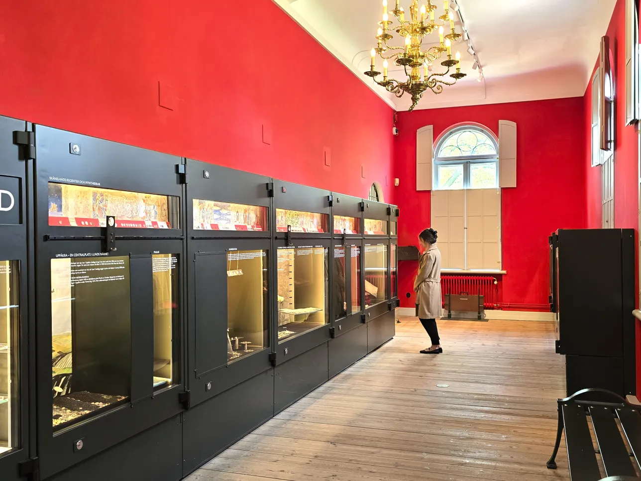 Ett avlångt rum med röda väggar, trägolv och ljuskrona i guld. En kvinna i beige kappa står och tittar in i en stor metallmonter som stäcker sig längs ena väggen.