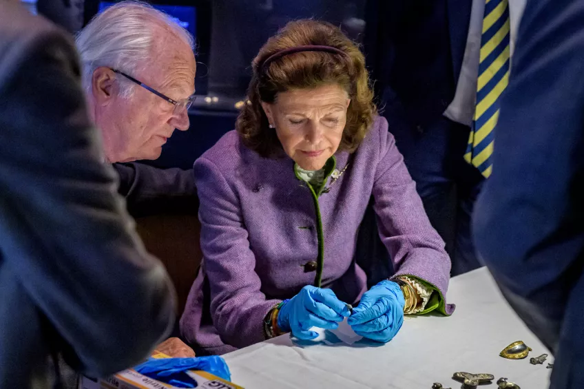 Bild på en man och en kvinna – Sveriges kung och drottning – som i blåa handskar undersöker arkeologiska fynd.